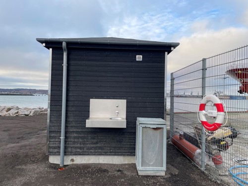 Toiletbygning med en håndvask på ydersiden, så lystfiskere kan rense fisk, samt en skraldespand placeret ved siden af bygningen